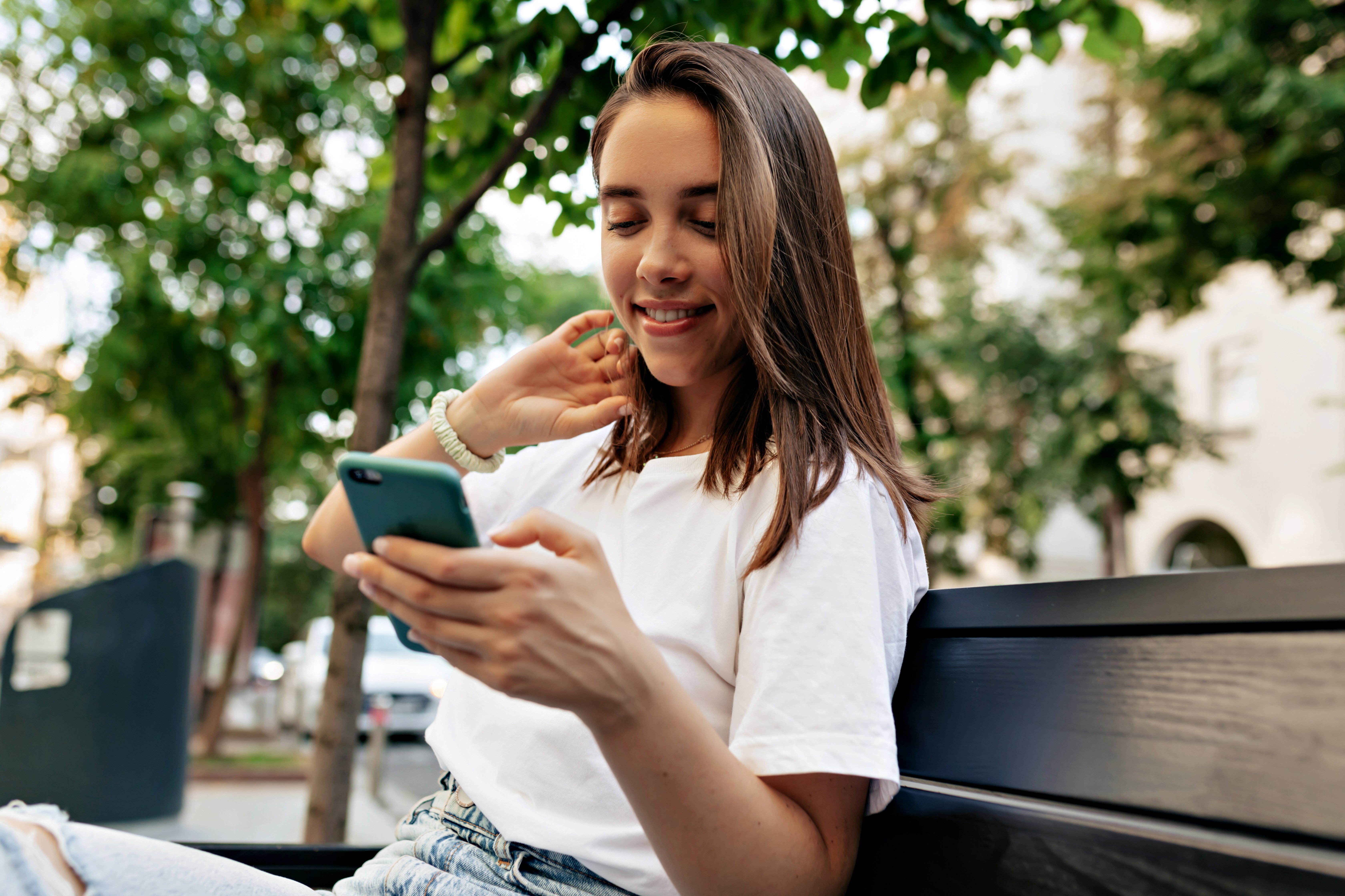 alluring-woman-with-loose-hair-white-short-is-touching-her-hair-looking-smartphone-while-sitting-city-bench-waiting-friends