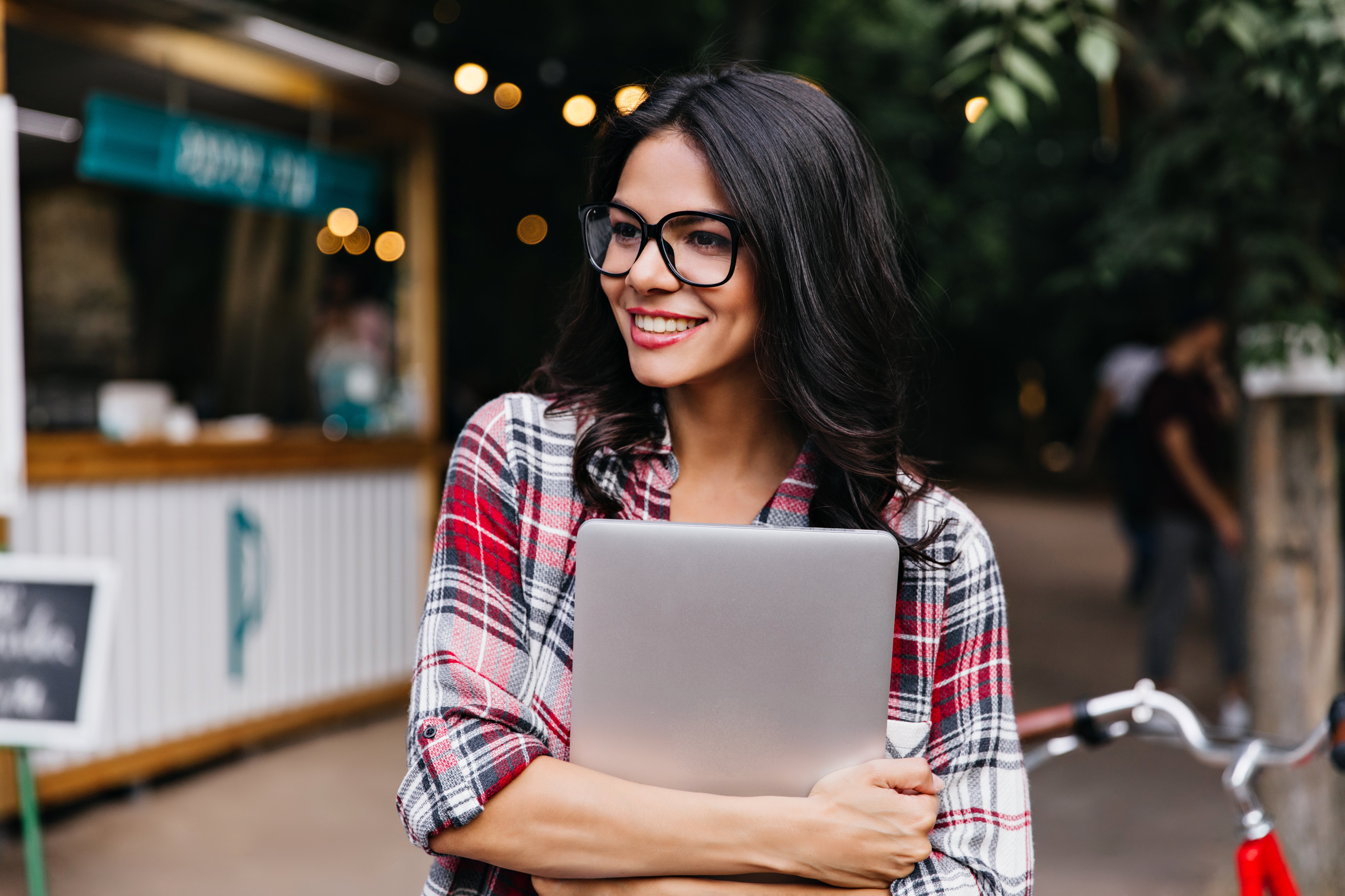 charming-female-student-with-wavy-hair-standing-on-the-street-pretty-tanned-girl-in-checkered-shirt-holding-laptop-and-looking-around
