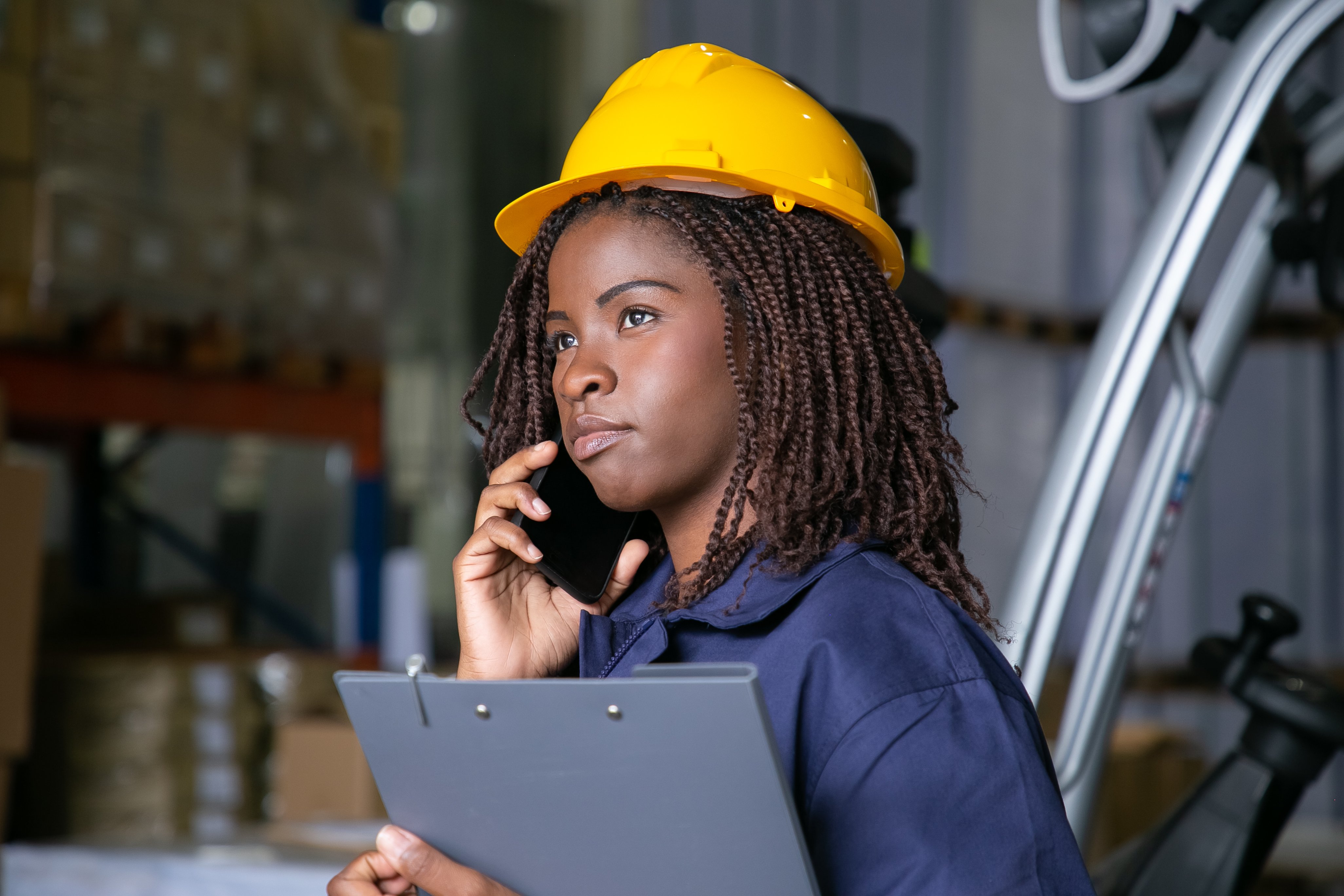 pensive-black-female-engineer-in-hardhat-standing-in-warehouse-and-talking-on-cellphone-shelves-with-goods-in-background-copy-space-labor-or-communication-concept