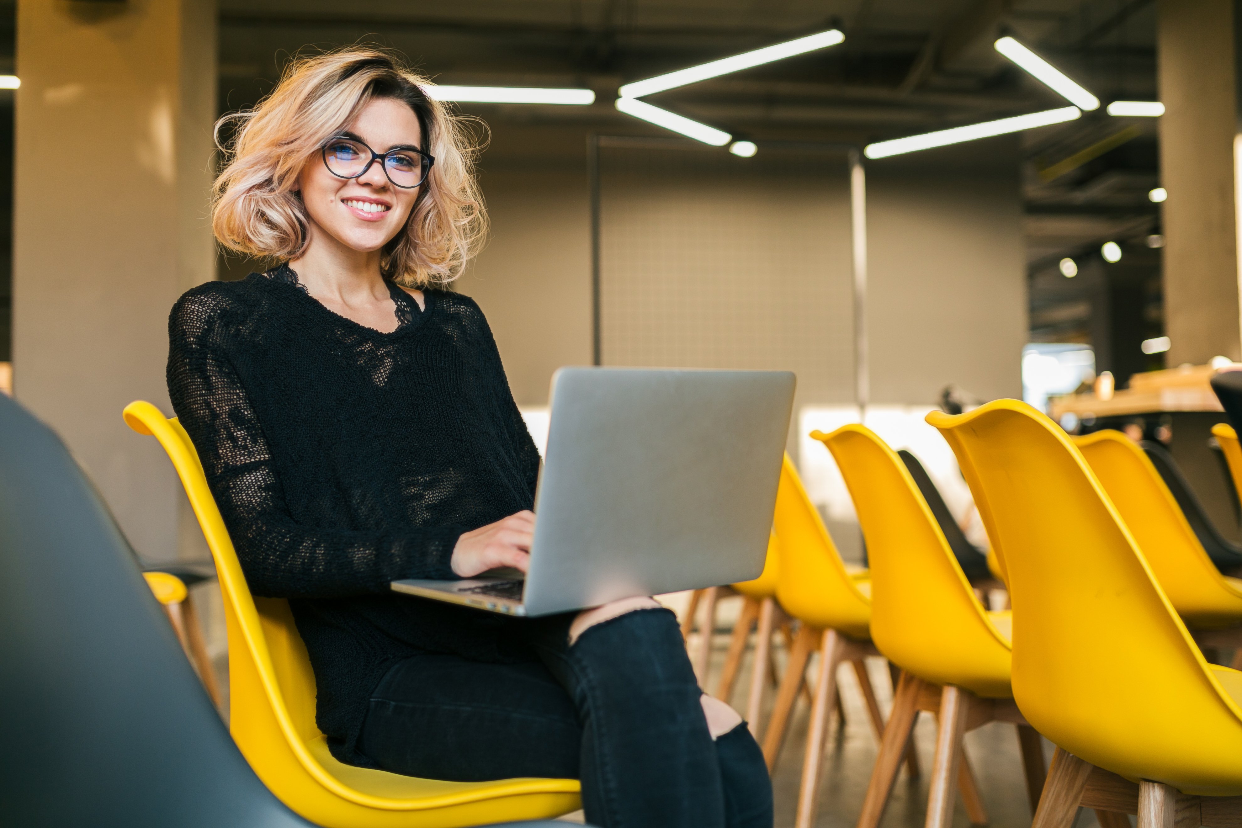 retrato-de-uma-jovem-mulher-atraente-sentada-na-sala-de-aula-trabalhando-no-laptop-de-oculos-o-aluno-aprendendo-na-sala-de-aula-com-muitas-cadeiras-amarelas