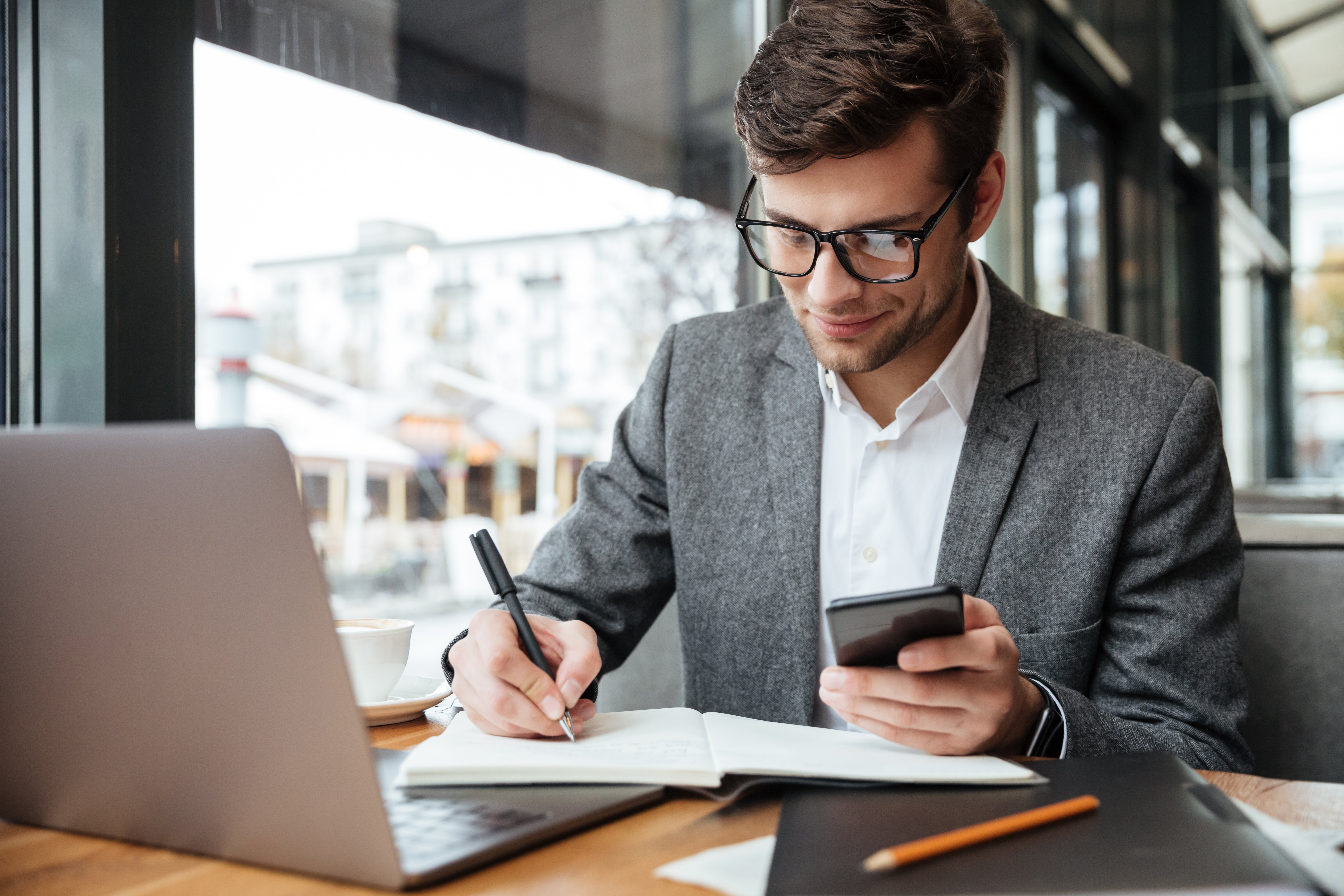 smiling-businessman-in-eyeglasses-sitting-by-the-table-in-cafe-with-laptop-computer-while-using-smartphone-and-writing-something