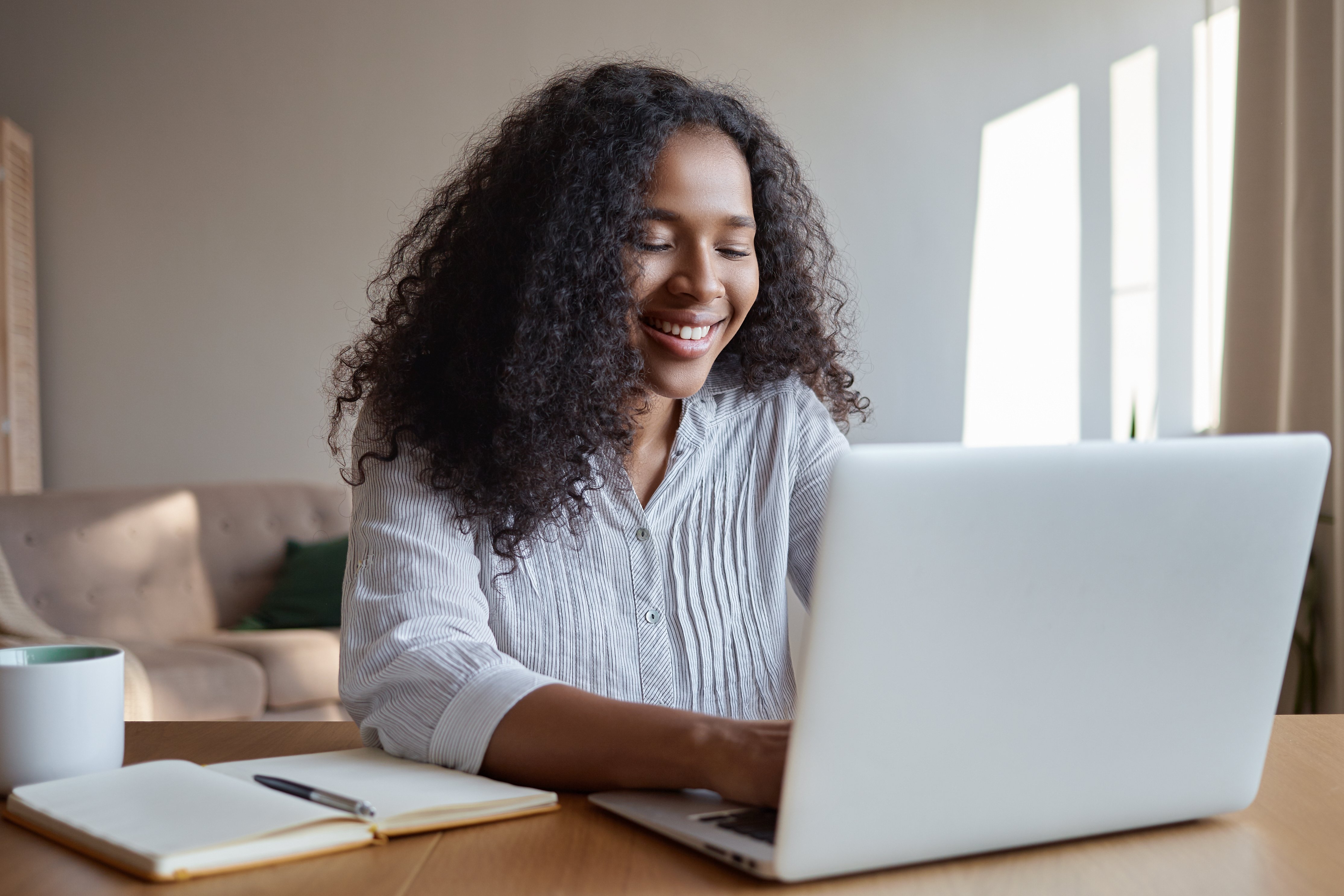 technology-occupation-freelance-and-networking-concept-charming-young-dark-skinned-female-marketing-expert-working-distantly-from-home-office-typing-on-laptop-making-notes-and-havning-coffee